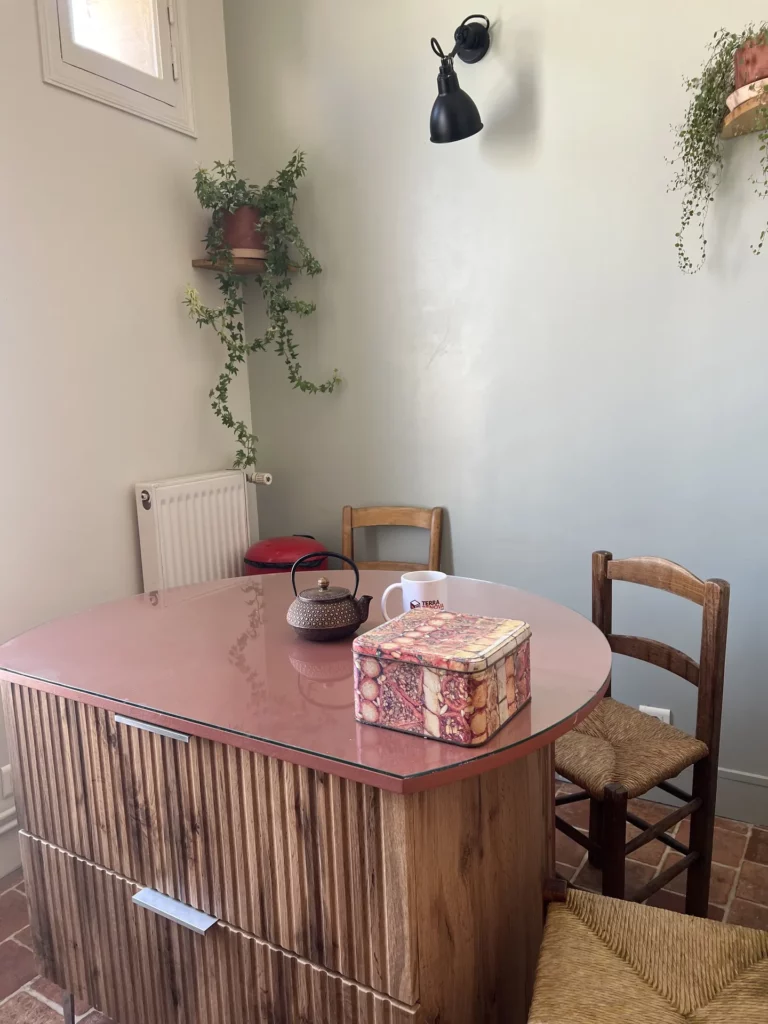 Coin repas d'une cuisine parisienne : alcôve peinte en vert Mizzle, meuble à tiroirs servant de table avec plateau arrondi sur mesure, chaises paillées et plantes suspendues